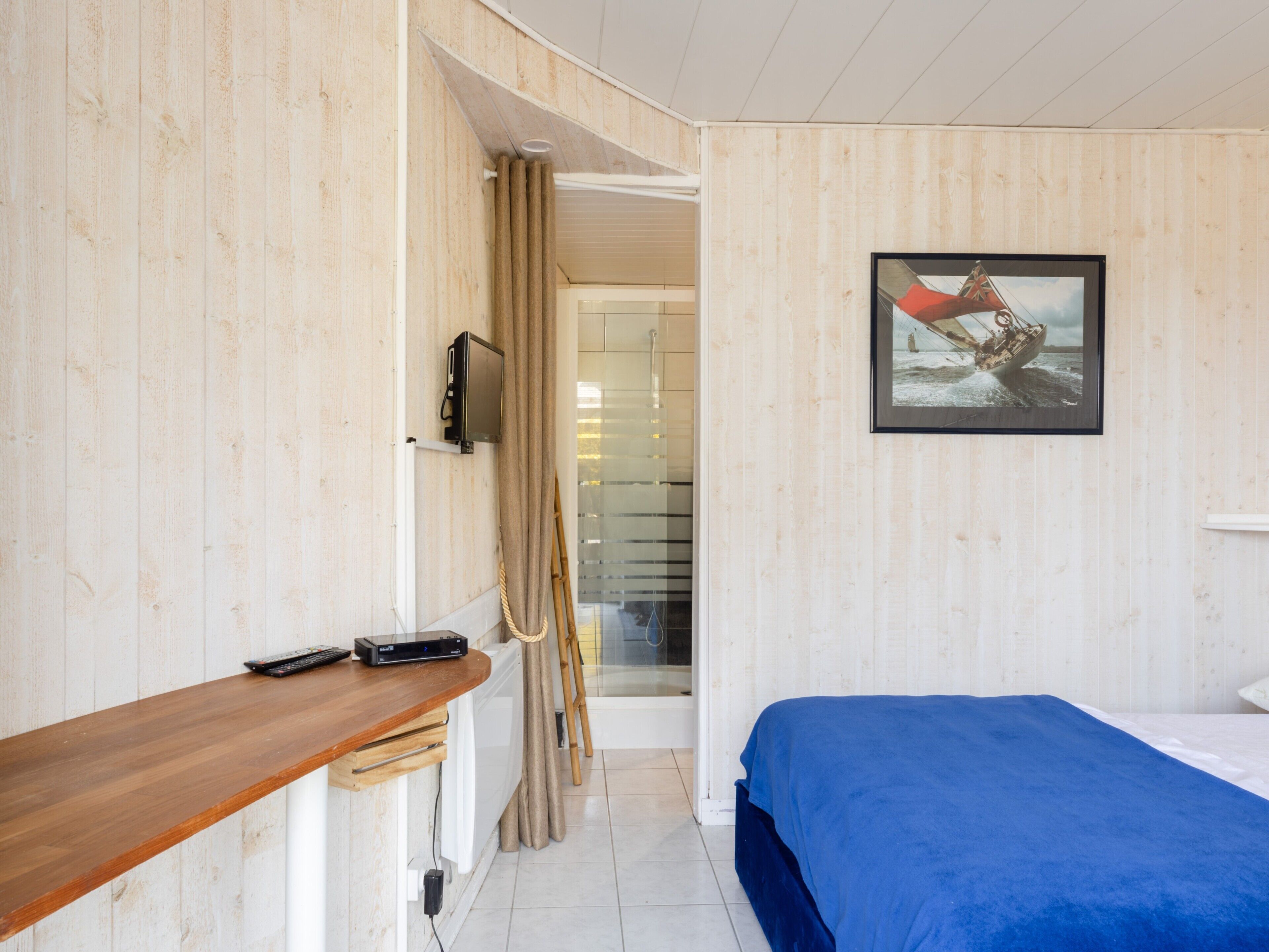 A room with a wooden wall, a bed with a blue bedspread, a TV mounted on the wall, and a remote control on a wooden table.