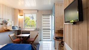 Ein kompaktes Zimmer mit einem Bett, einem Esstisch mit Stühlen, einer Küchenzeile inklusive Mikrowelle, einem an der Wand befestigten Fernseher und einem Fenster mit Jalousien.
