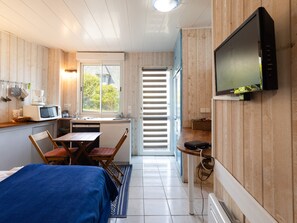 A compact room with a bed, a dining table with chairs, a kitchenette with a microwave, a television mounted on the wall, and a window with blinds. - Cote de granit rose, vue mer, studio ground floor renovated this year (Perros-Guirec)