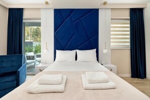 Una habitación de hotel moderna con una cama grande, dos mesitas de noche, un sillón azul y vistas al exterior.