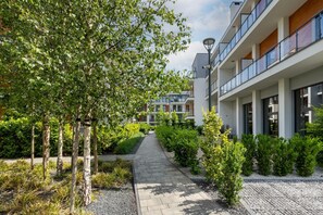 Ein Weg führt zu einem modernen Gebäude mit Balkonen und einem Garten mit Bäumen und Sträuchern.