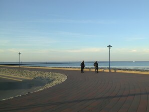 A paved walkway along a coastal area with two individuals standing, a railing, and a lamp post. - Deichblick Apartment in Beautiful Beach Location with Wi-Fi (Neuharlingersiel)