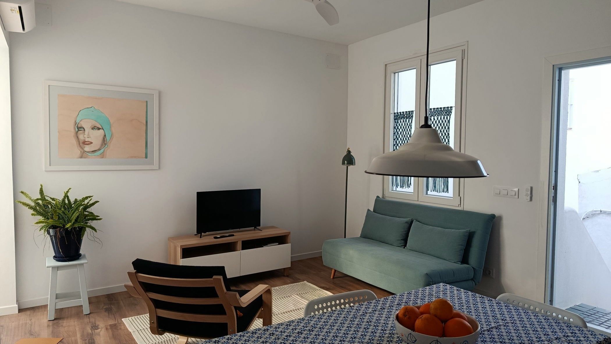 A living room with a sofa, a television, a dining table with a bowl of fruit, and a potted plant.