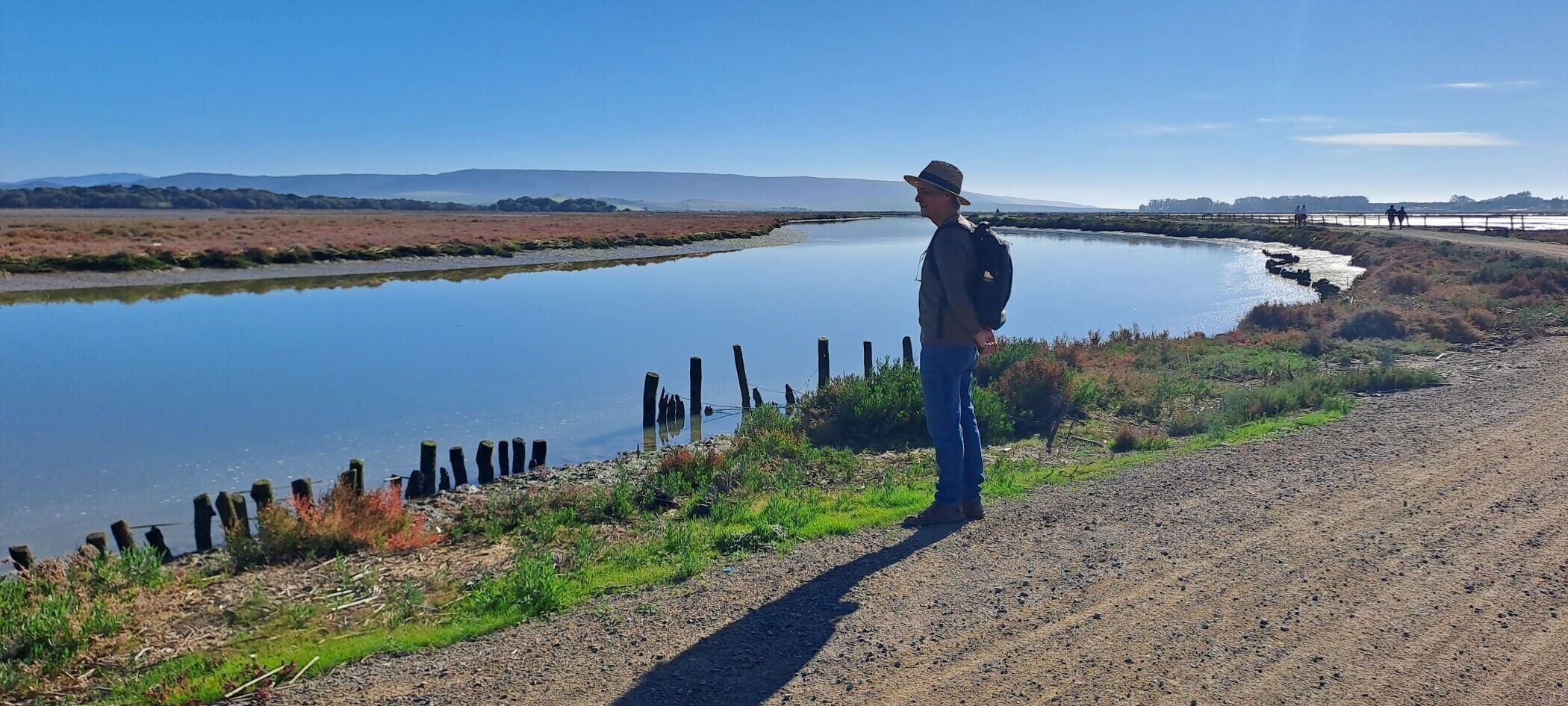 A person standing by a calm river with a fence and distant hills.