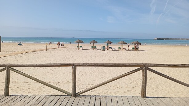 Una playa con un muelle de madera, sillas de playa y sombrillas de paja.