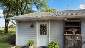 Ein Haus mit einer weißen Tür, einer Veranda mit zwei Blumentöpfen, einem Holzkabinett mit der Texas-Landesflagge und einem Metalldach.