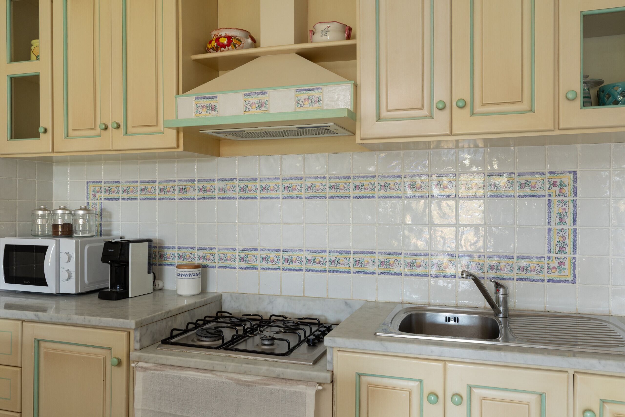 Una cocina con microondas, tostadora, estufa a gas, pileta y azulejos decorativos.