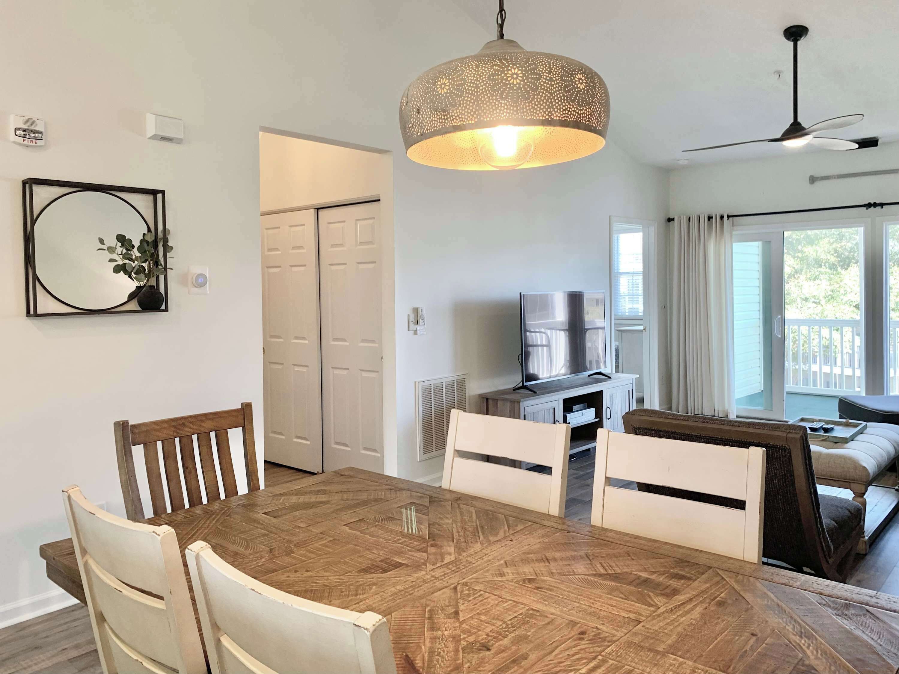 A dining area with a wooden table and chairs, a hanging light fixture, a television on a stand, and a sliding glass door leading to an outdoor area.