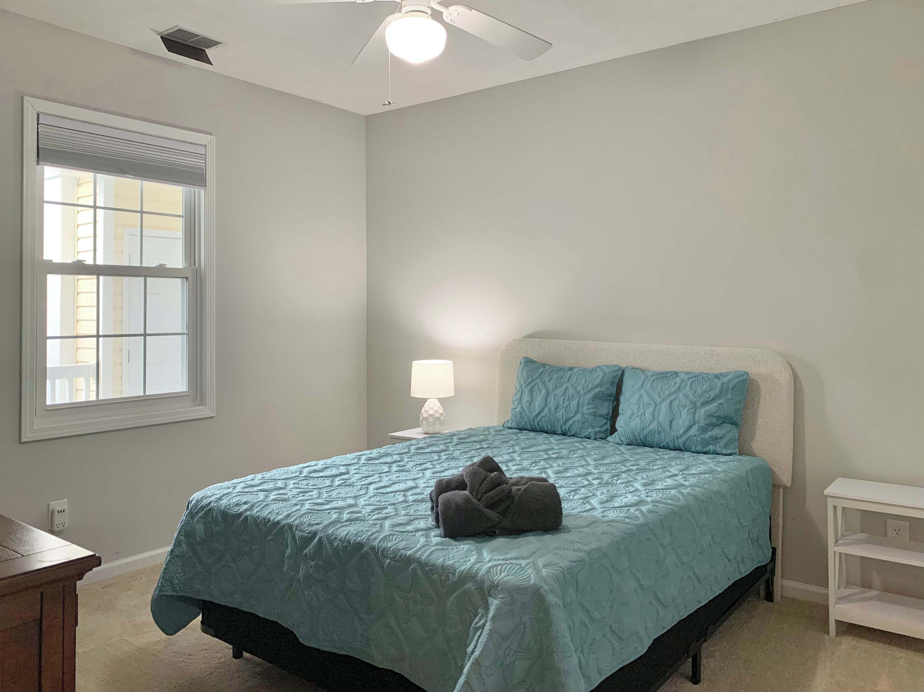 Une chambre à coucher avec un lit, une table de chevet, une lampe, une fenêtre et un ventilateur de plafond.