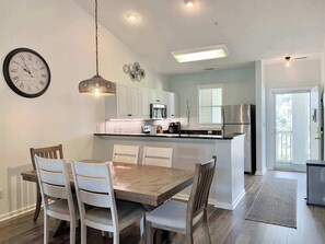 A modern kitchen with white cabinets, a central island, a dining table with chairs, and a clock on the wall. - Peaceful 2Bed/2Bath Condo | King Bed | Pool View (North Myrtle Beach)