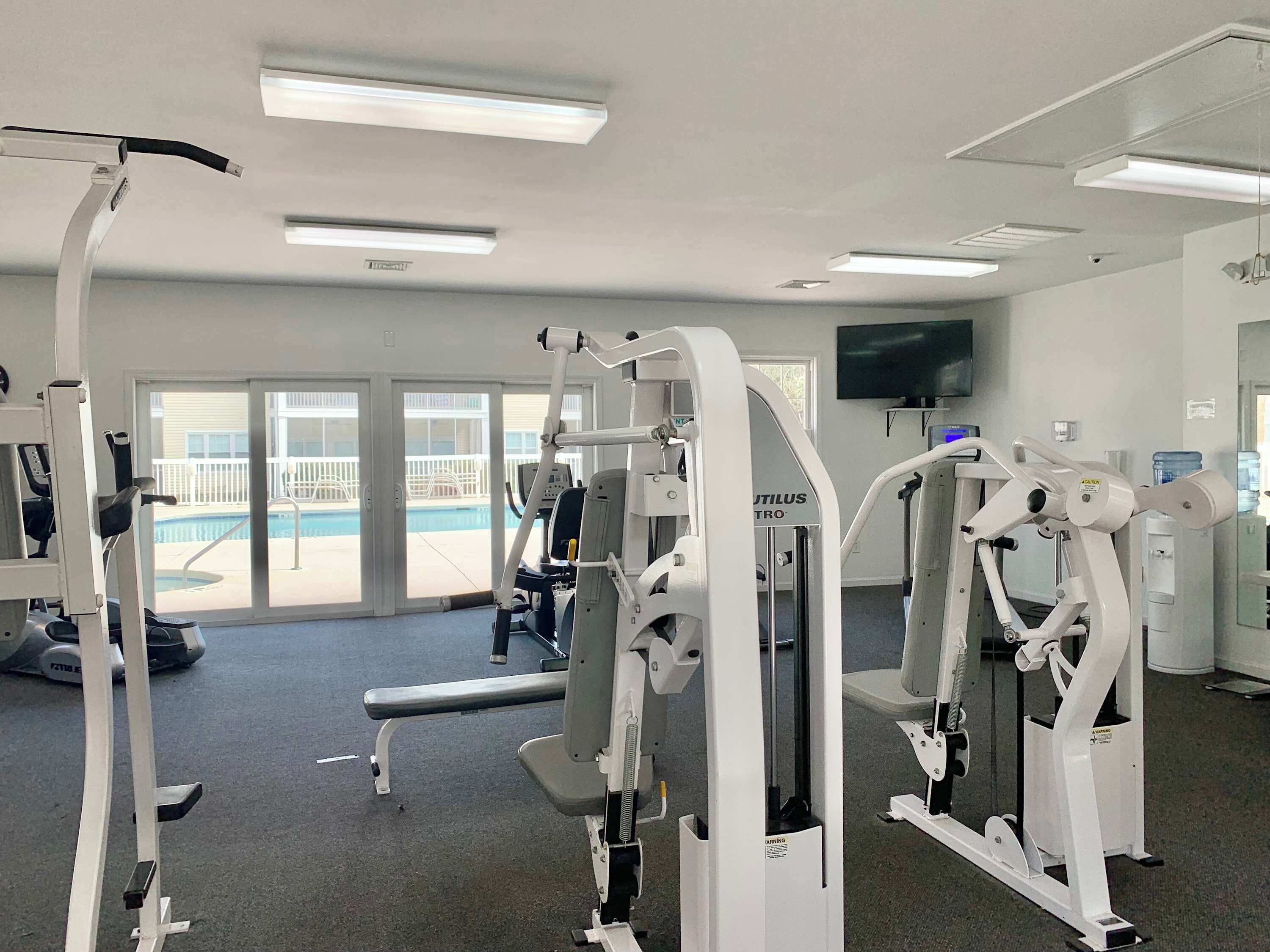 A gym with various exercise machines, a pool view, and a TV on the wall.