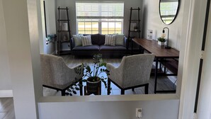 A room with a dark-colored sofa, two chairs, a wooden table, a mirror, and a window with blinds. - Beach Cottage: Cozy I Clean I Family I Beaches (Port St. Lucie)