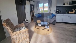 Een gezellig, klein leefruimte met een bank, een rieten fauteuil, een salontafel, een keukenhoek en uitzicht door het raam.