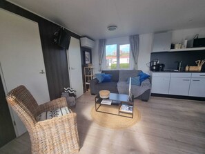 Ein gemütlicher Wohnraum mit einer Couch, einem Korbsessel, einem Couchtisch, einer Küchenzeile und Blick durch das Fenster.