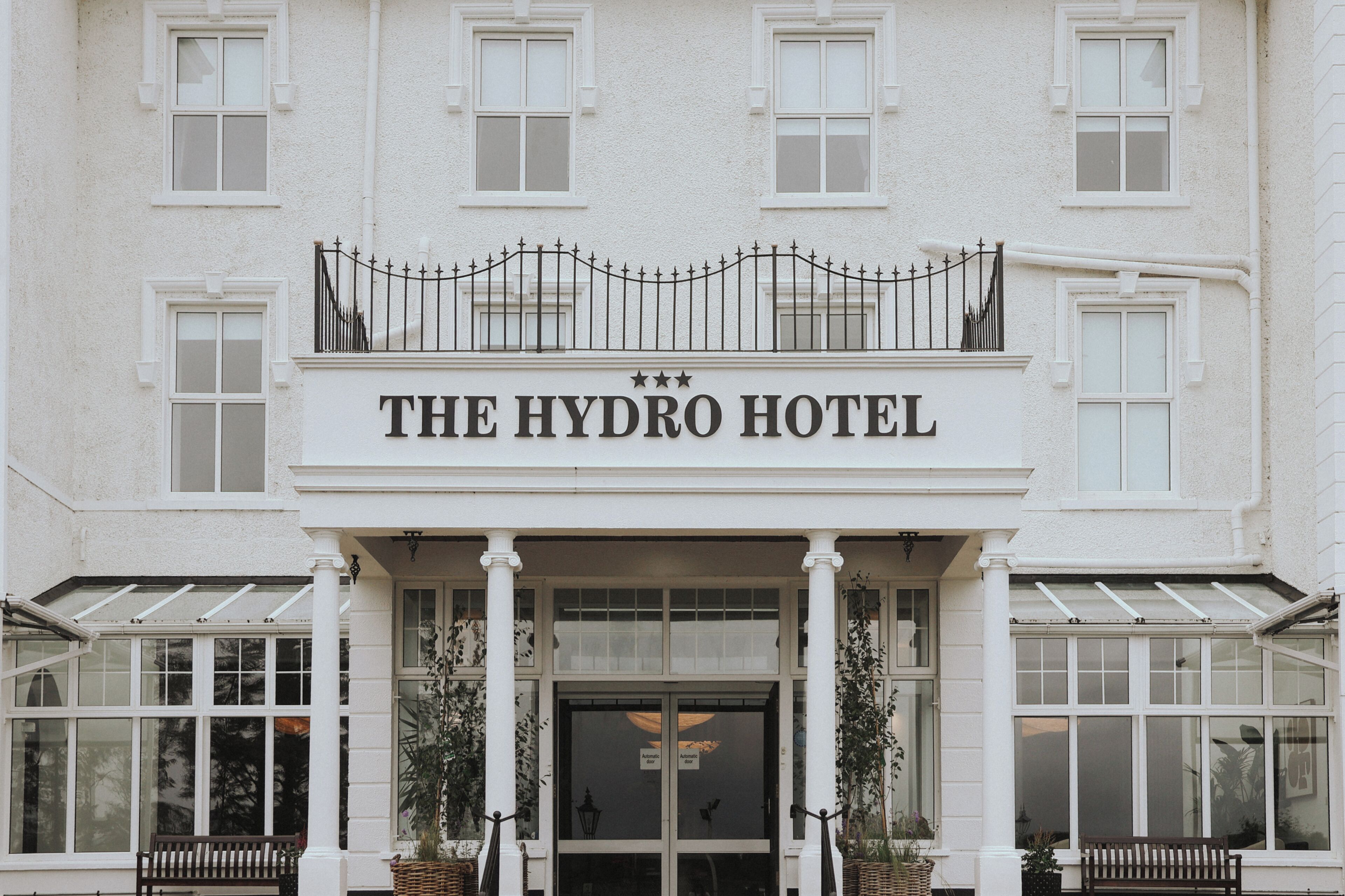 A building with a sign reading 'THE HYDRO HOTEL' above the entrance, featuring a balcony with a wrought iron railing, large windows, and potted plants.