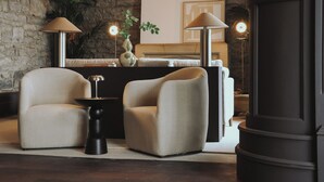 A modern room with two armchairs, a small side table, a lamp, a plant, and a bed with a headboard.