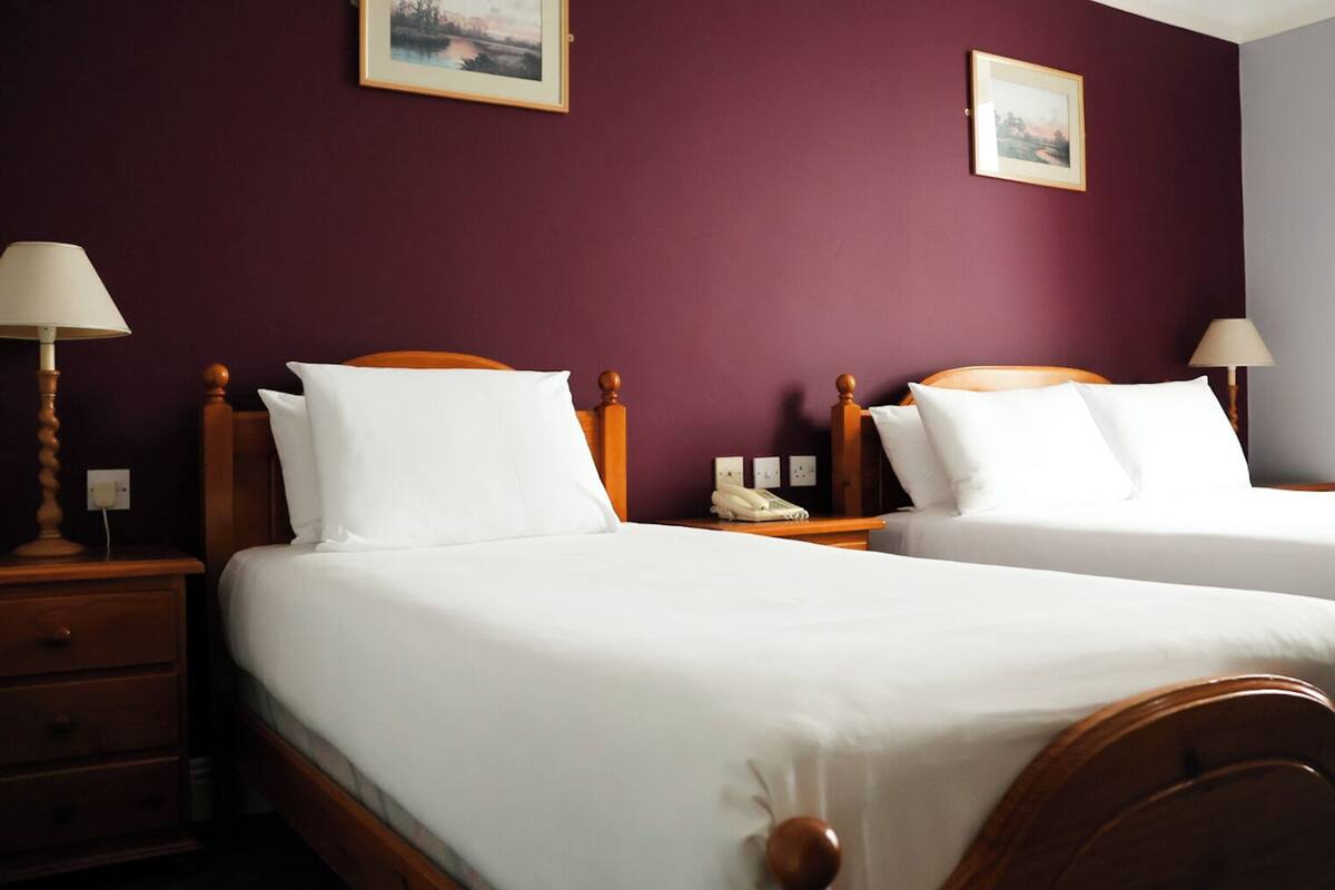 A hotel room with two wooden beds, white bedding, a nightstand with a lamp, a phone, and two framed pictures on the wall.