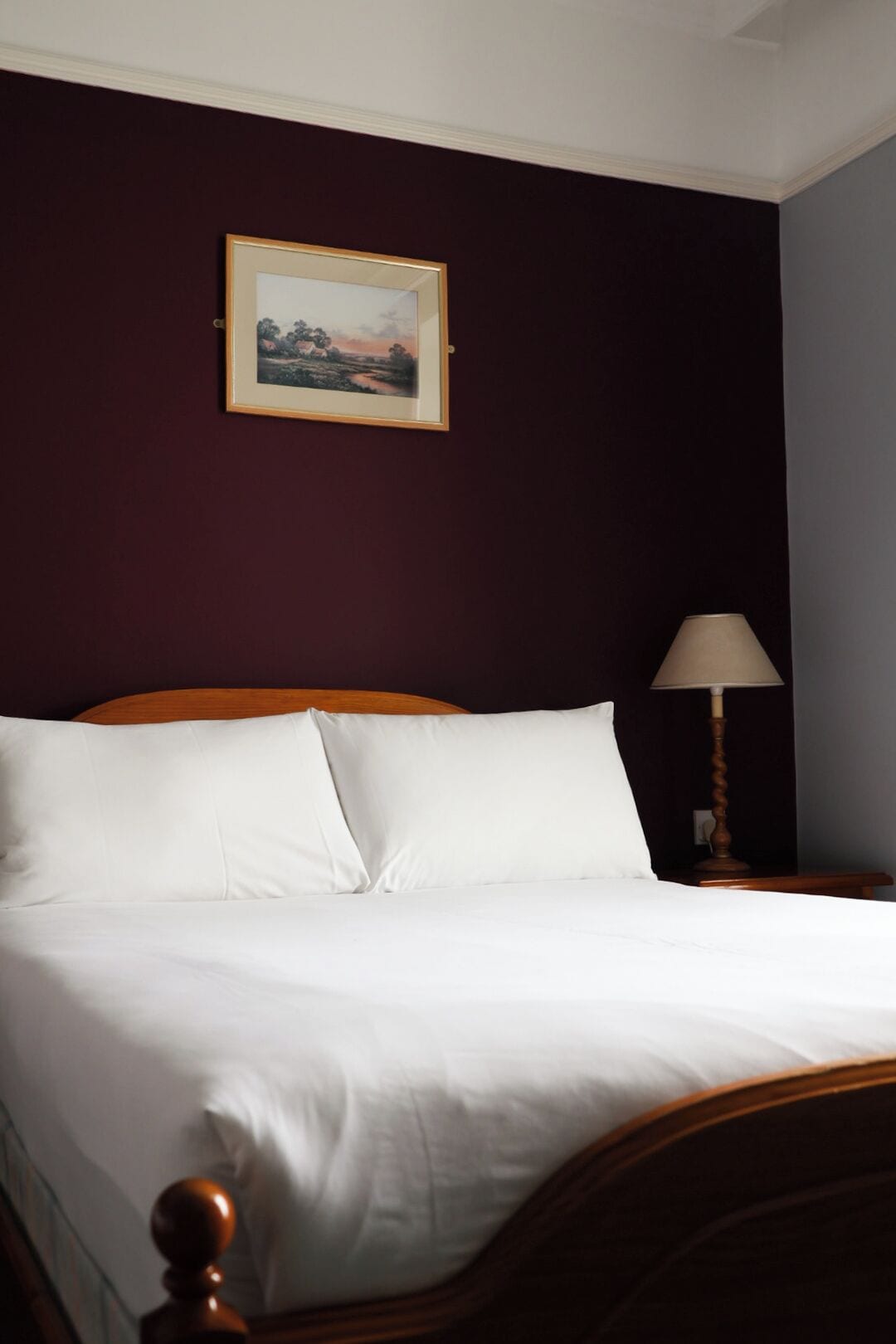 Una cama bien hecha con sábanas blancas, cabecera de madera, un cuadro en la pared y una lámpara de noche.