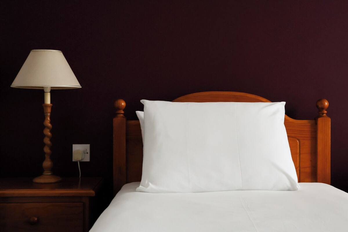 A neatly made bed with a wooden headboard, a white pillow, and a lamp on a wooden nightstand.