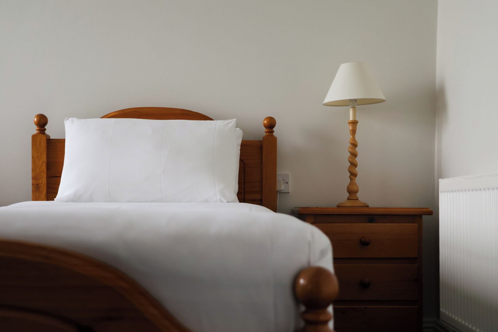 A neatly made bed with white linens, a wooden headboard, a bedside table with a lamp, and a drawer.