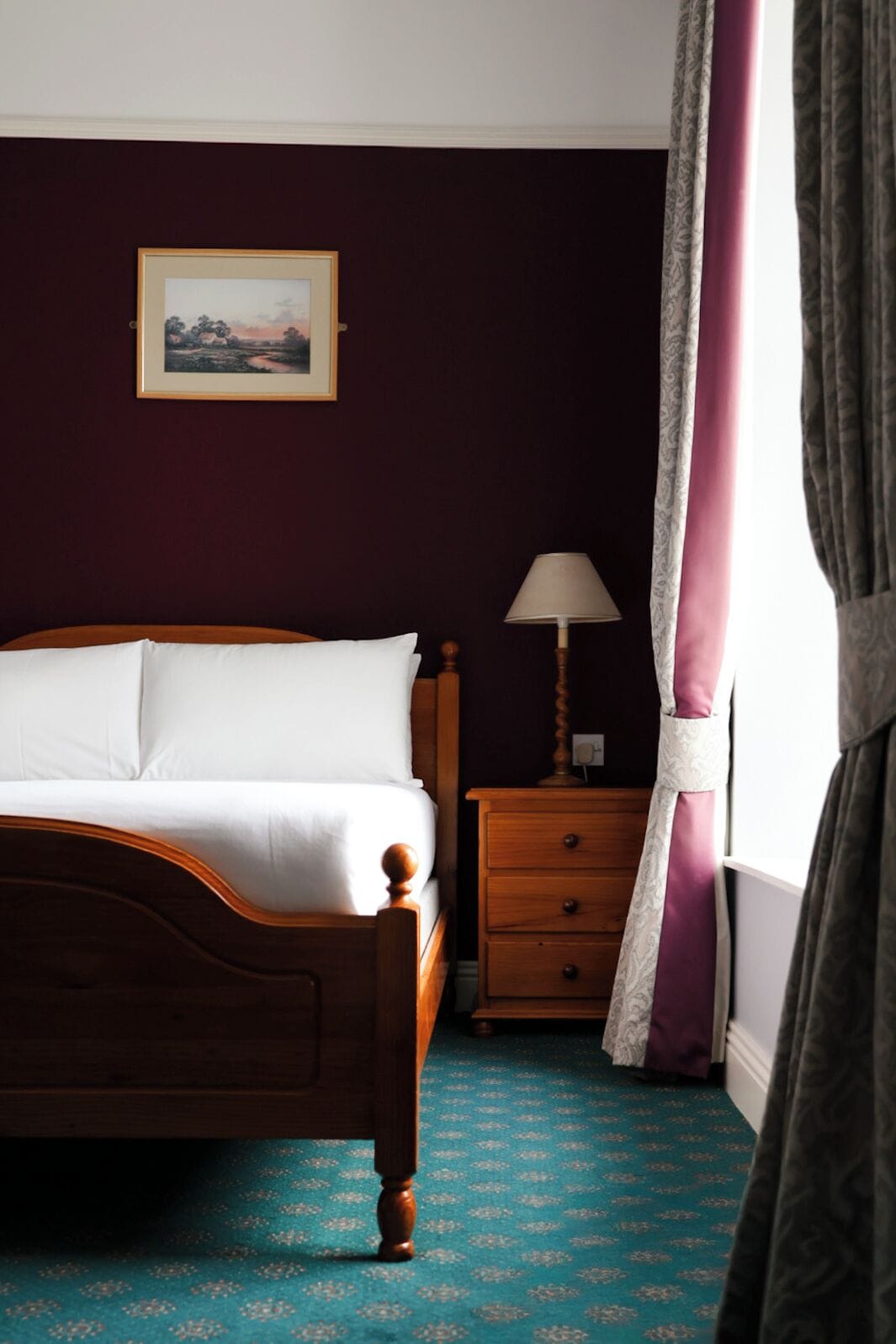 A bedroom with a wooden bed, a nightstand with a lamp, a wall-mounted picture, and a window with curtains.
