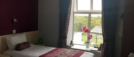 A hotel room with two beds, a window with curtains, a small table with a plant, and a view of greenery outside.