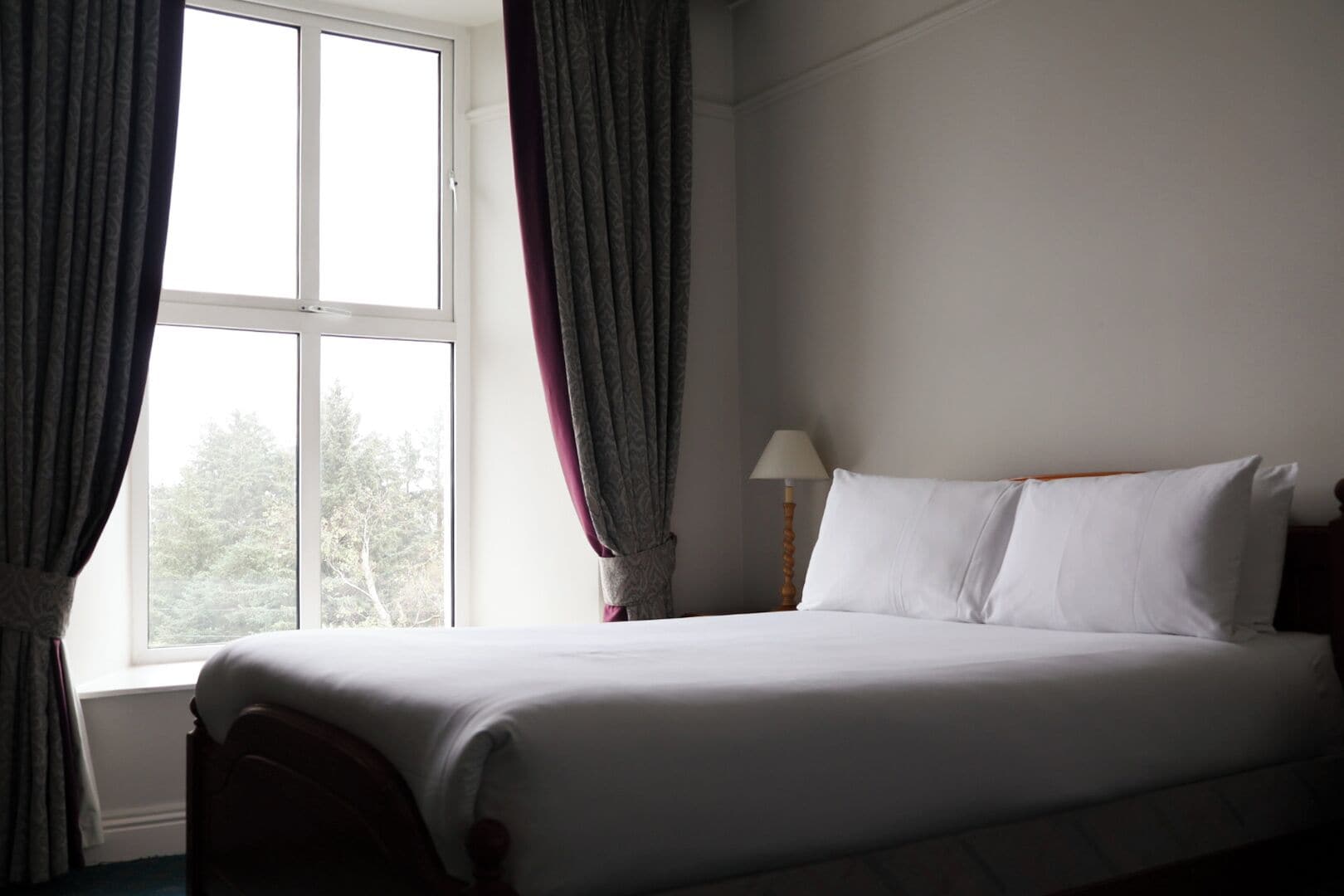A hotel room with a bed, curtains, a lamp, and a window.