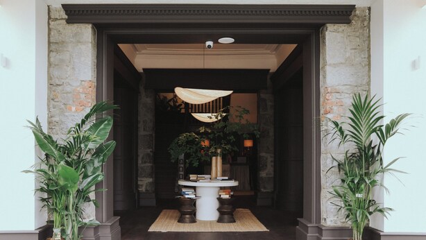 A lobby with a central table surrounded by potted plants, a decorative hanging light, and a stone wall.