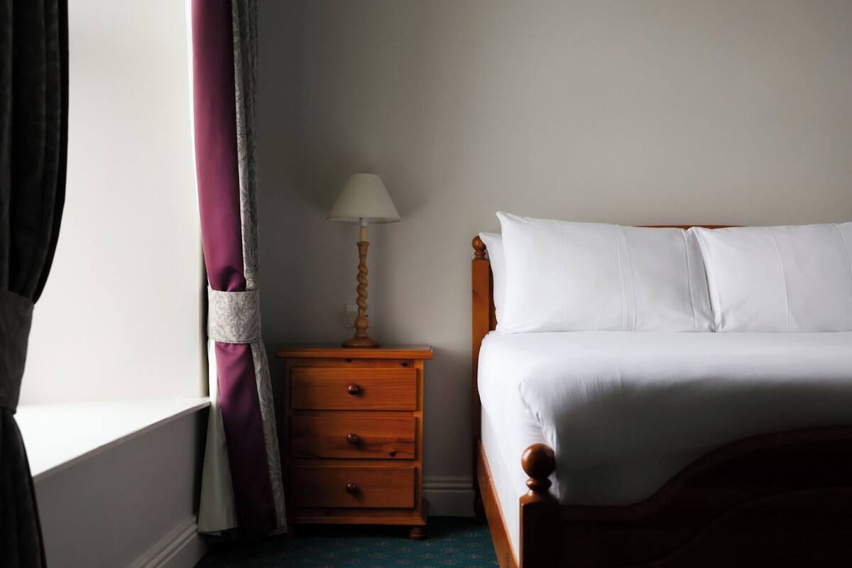 A bedroom with a bed, a wooden nightstand with a lamp, and a window with curtains.