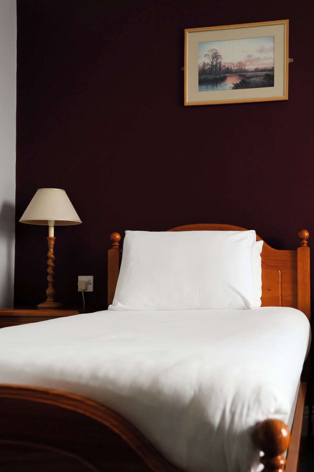 Una cama con ropa de cama blanca, un cabecero de madera, una lámpara de noche y un cuadro en la pared.