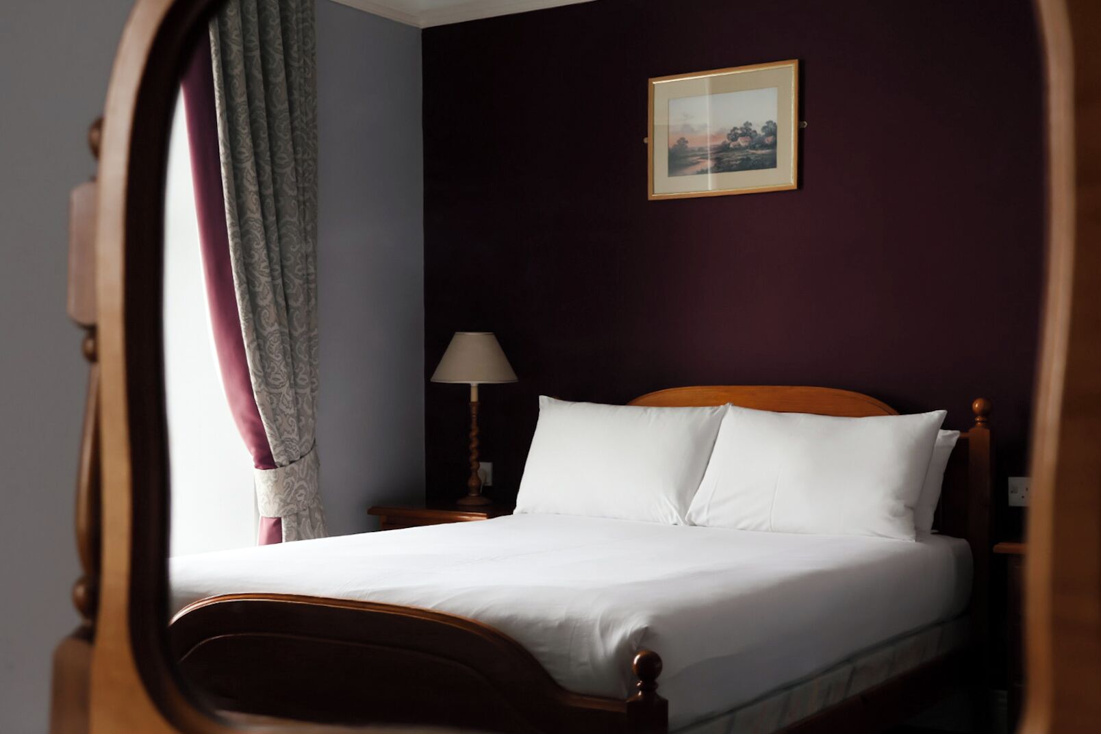 A neatly made bed with white linens in a room with dark walls, a framed picture on the wall, and a lamp on a bedside table.