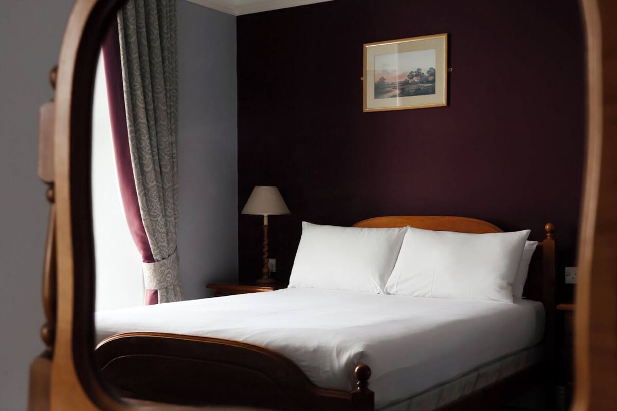 A neatly made bed with white linens in a room with dark walls, a framed picture on the wall, and a lamp on a bedside table.