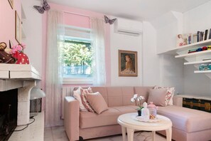 A living room with a pink sofa, a small round table, a fireplace, a window with curtains, and a bookshelf.