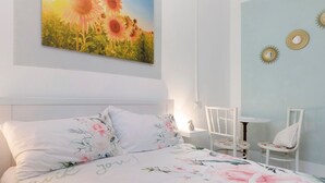 Une chambre à coucher avec un lit, des coussins à motifs floraux, un tableau représentant des tournesols accroché au mur, et une petite table avec des chaises.
