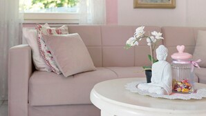 Un canapé rose agrémenté d’un coussin décoratif, une petite statue de Bouddha, un bocal en verre avec un couvercle rose et une table blanche sur laquelle repose une nappe en dentelle.