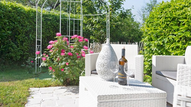 Un jardin agrémenté d’un espace extérieur avec des sièges blancs, d’une treille en métal et de fleurs roses.