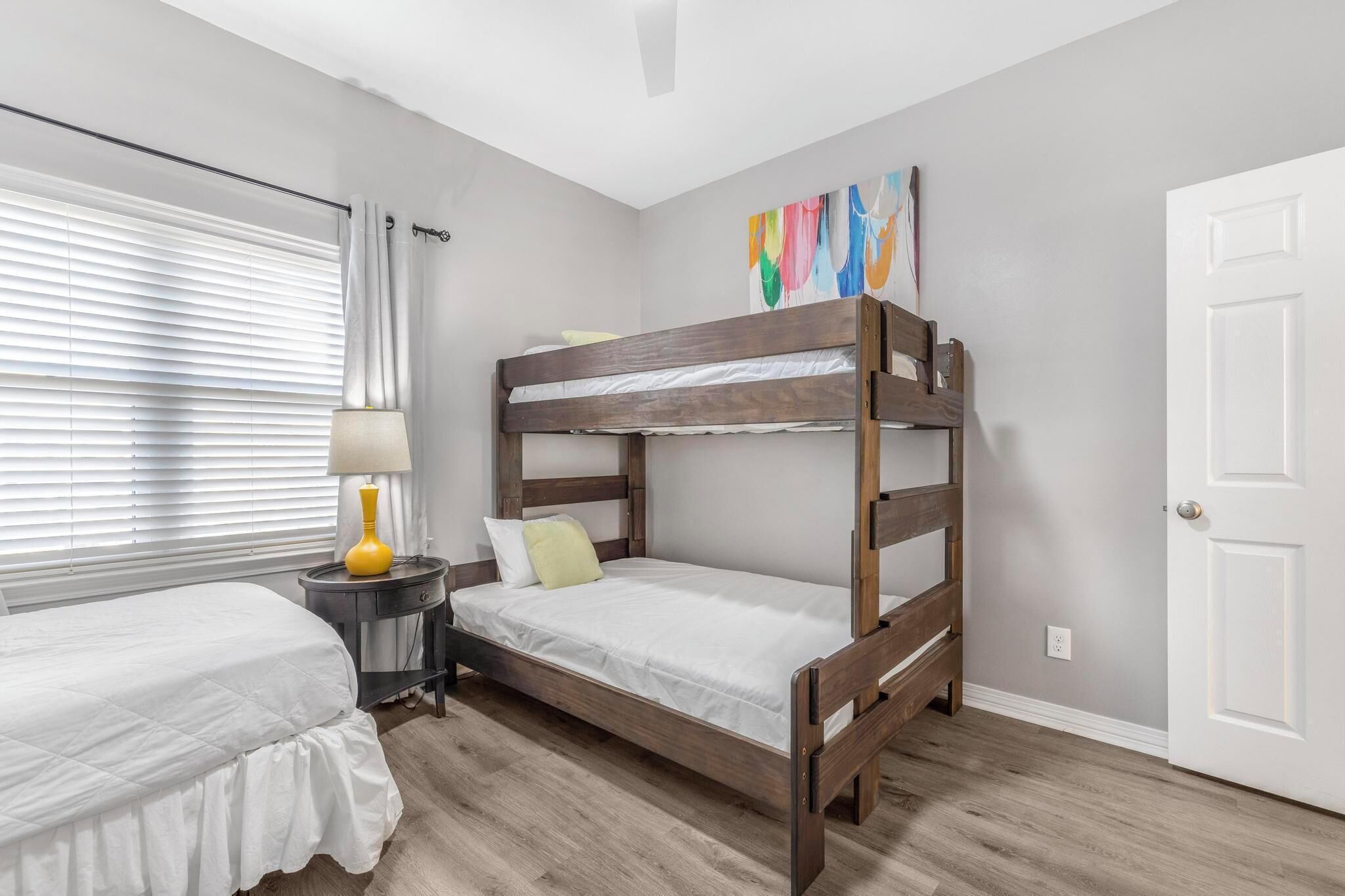 A bedroom with a bunk bed, a bedside table with a lamp, a window with blinds, and a door.