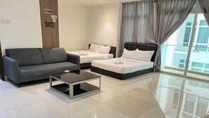 Quarto de hotel moderno com sofá cinza, duas camas de solteiro, mesa de centro de vidro e uma janela grande com cortinas.