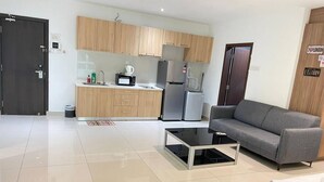 Uma sala de estar moderna com um sofá cinzento, uma mesa de centro preta e uma área de cozinha com armários de madeira e eletrodomésticos.