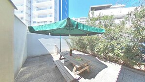 Um pátio com mesa de madeira e guarda-sol verde, cercado por plantas e edifícios.