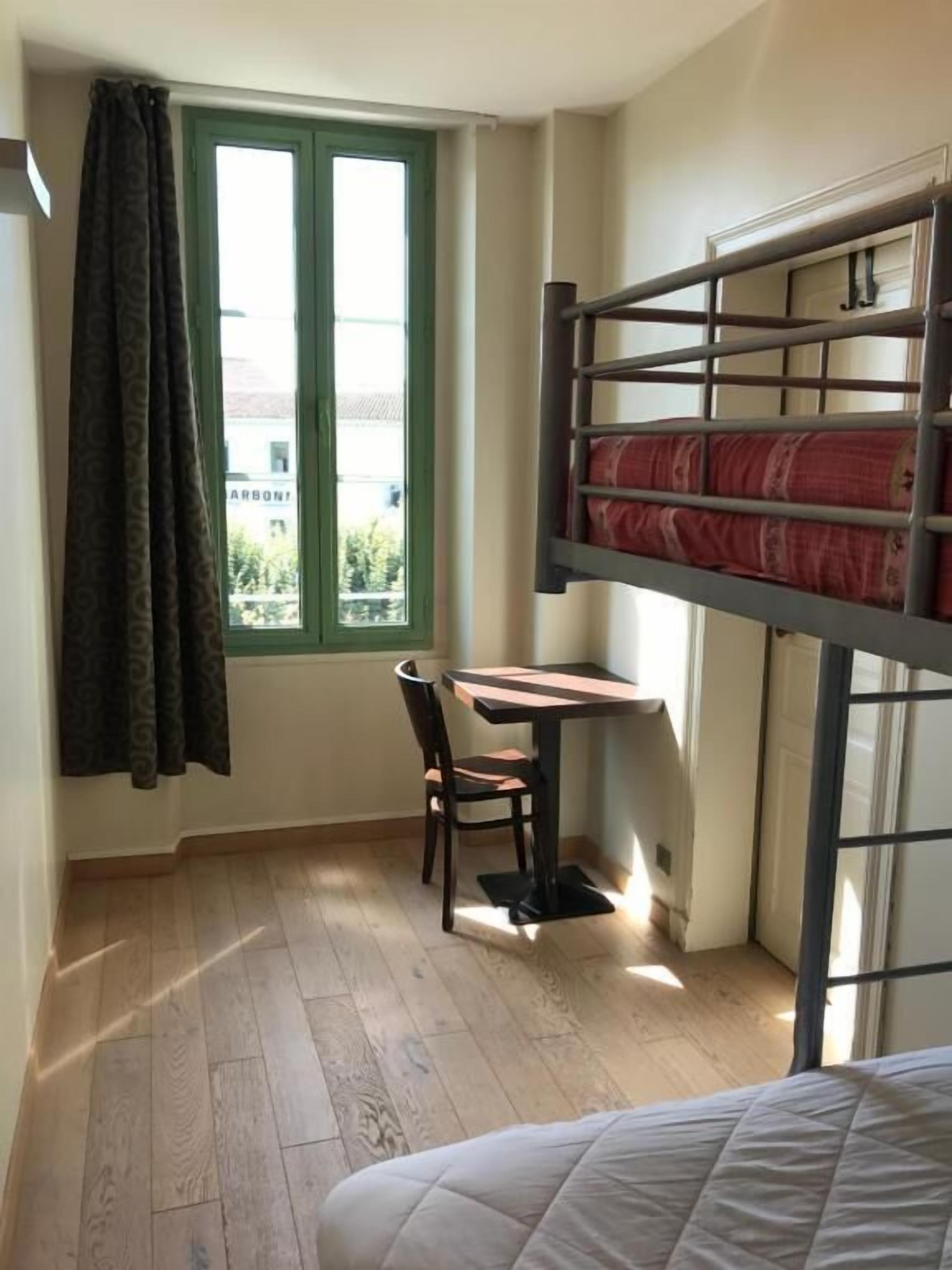 A dormitory room with a bunk bed, a desk with a chair, a window with curtains, and a view of buildings outside.