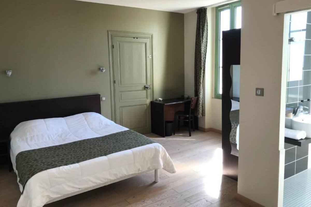 Un dormitorio con cama, escritorio, silla, puerta y baño con lavamanos y espejo.