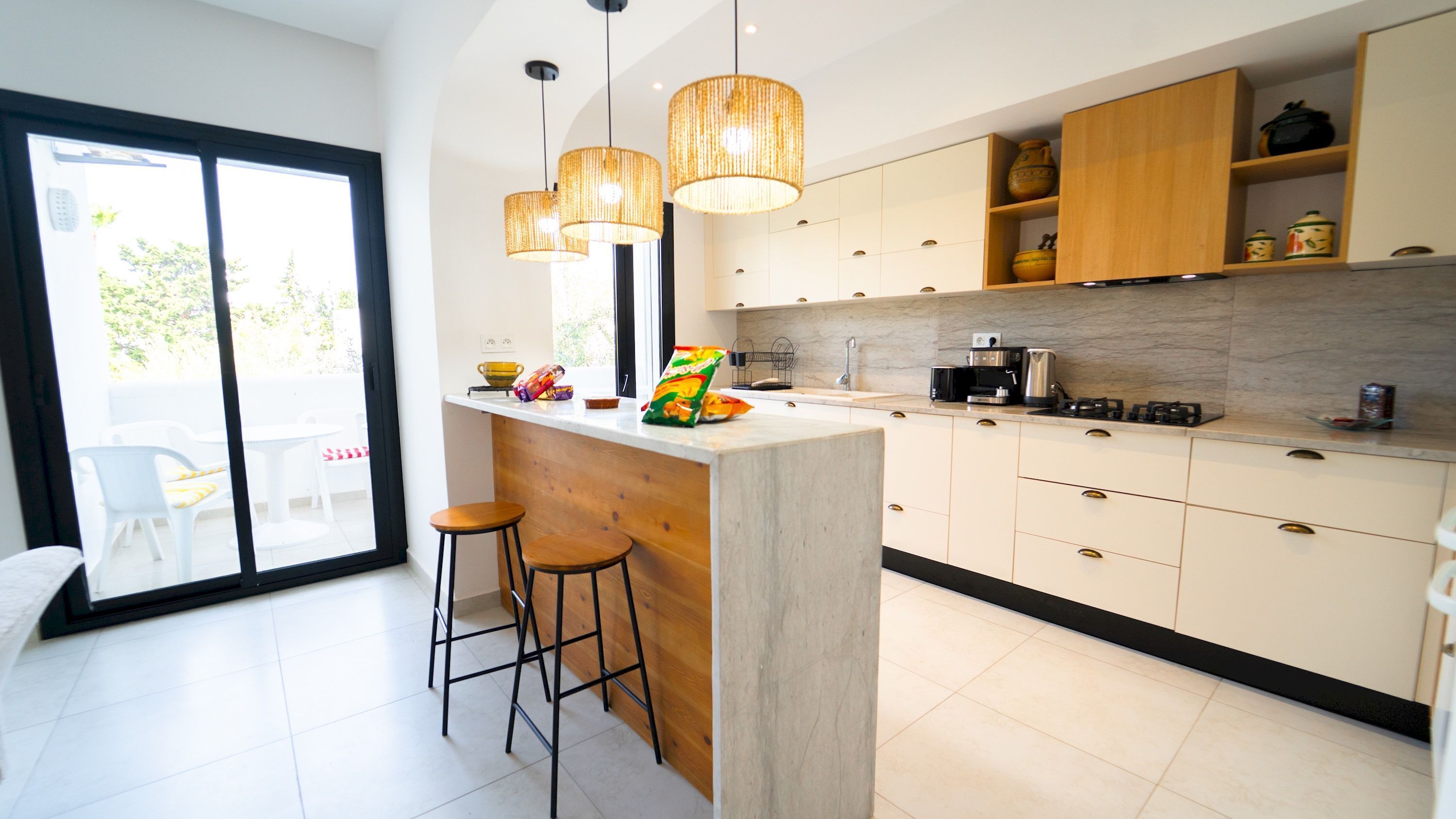 A modern kitchen with a central island, bar stools, pendant lights, and a view to a balcony.