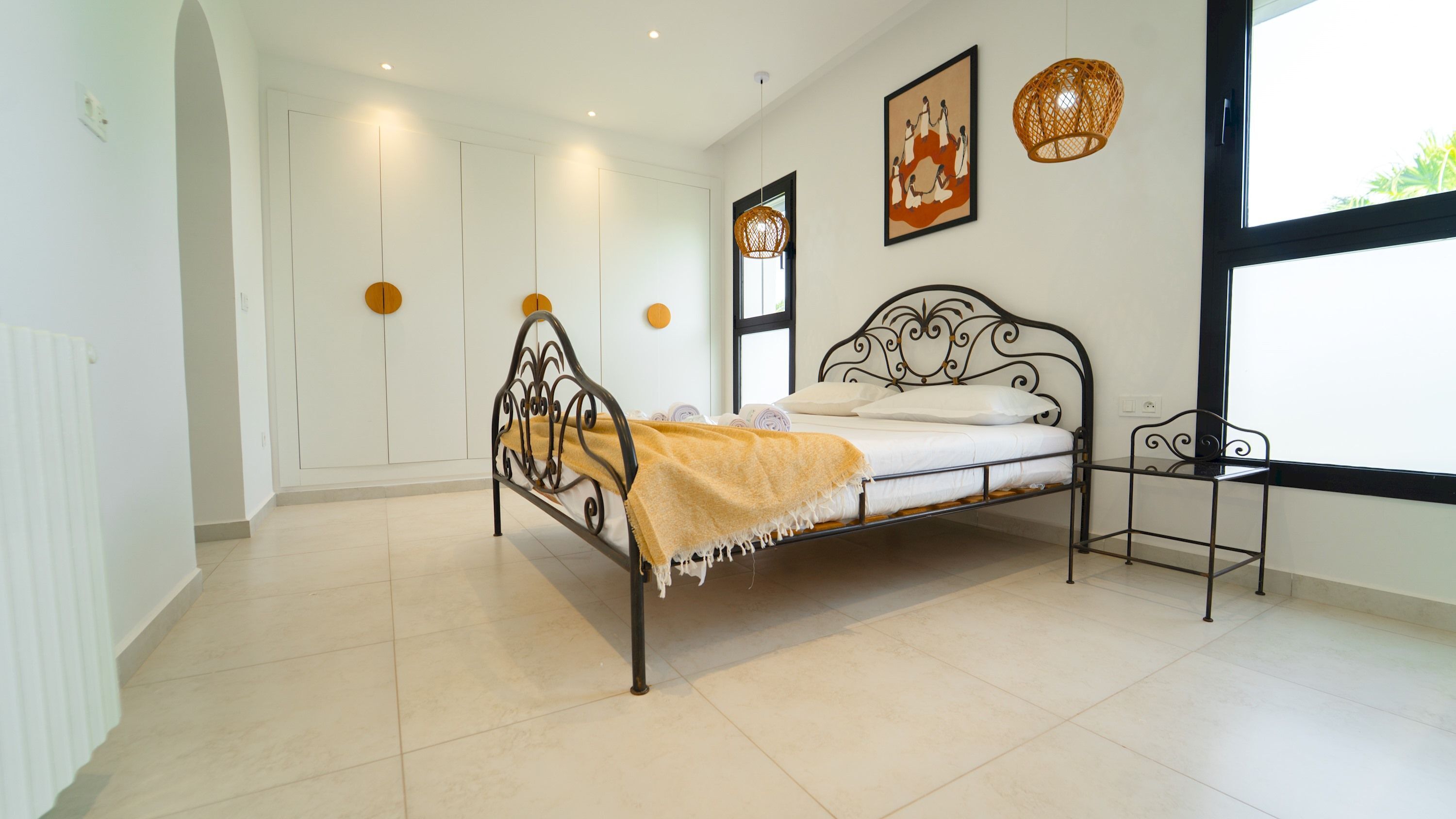 Une chambre avec un lit en métal, un matelas blanc, un plaid jaune et une petite table de chevet. Une fenêtre offre une vue sur la verdure à l’extérieur.