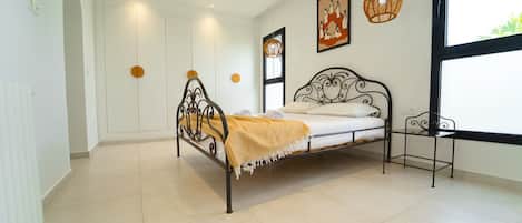A bedroom with a metal bed frame, a white mattress, a yellow throw blanket, and a small bedside table. There is a window with a view of greenery outside.