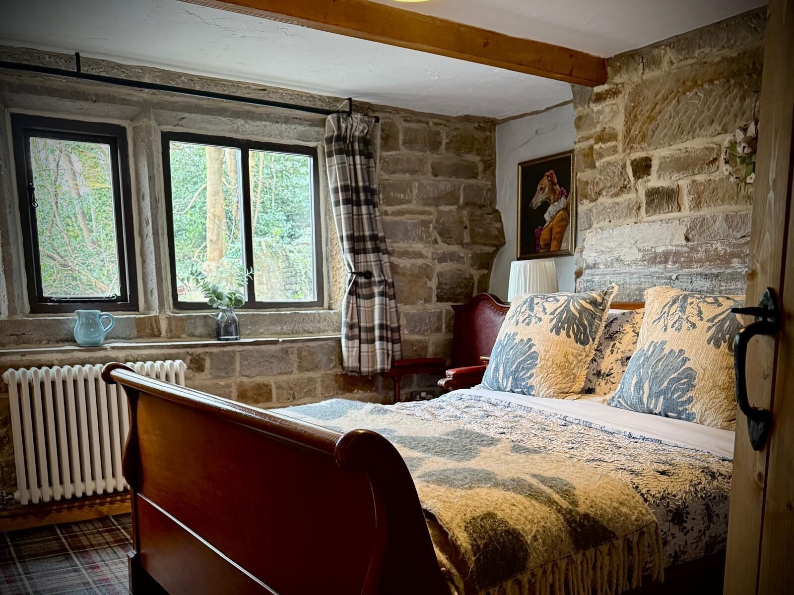 A rustic bedroom with a wooden bed, stone walls, a radiator, and a window with a view of trees.