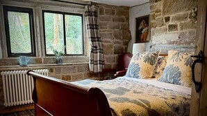 Ein rustikales Schlafzimmer mit einem hölzernen Bett, Steinwänden, einer Heizung und einem Fenster mit Blick auf Bäume.