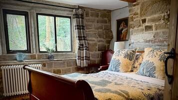 A rustic bedroom with a wooden bed, stone walls, a radiator, and a window with a view of trees.