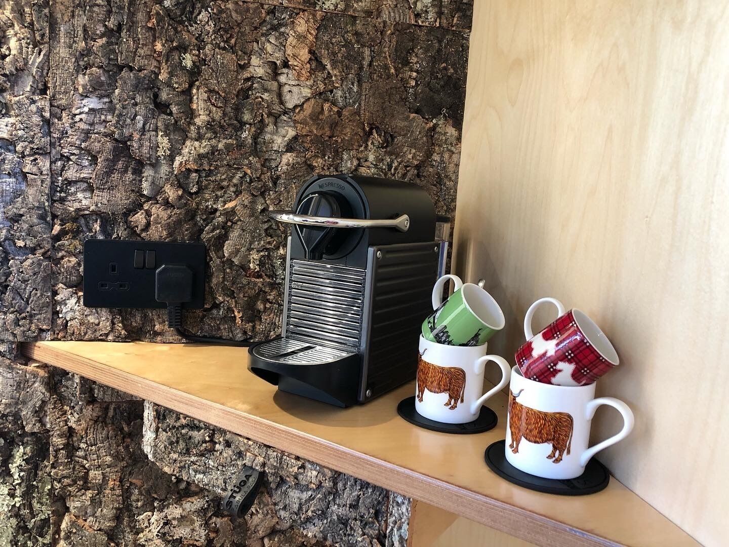 Una cafetera sobre una estantería, dos tazas con diseños de animales y una pared de piedra con textura.