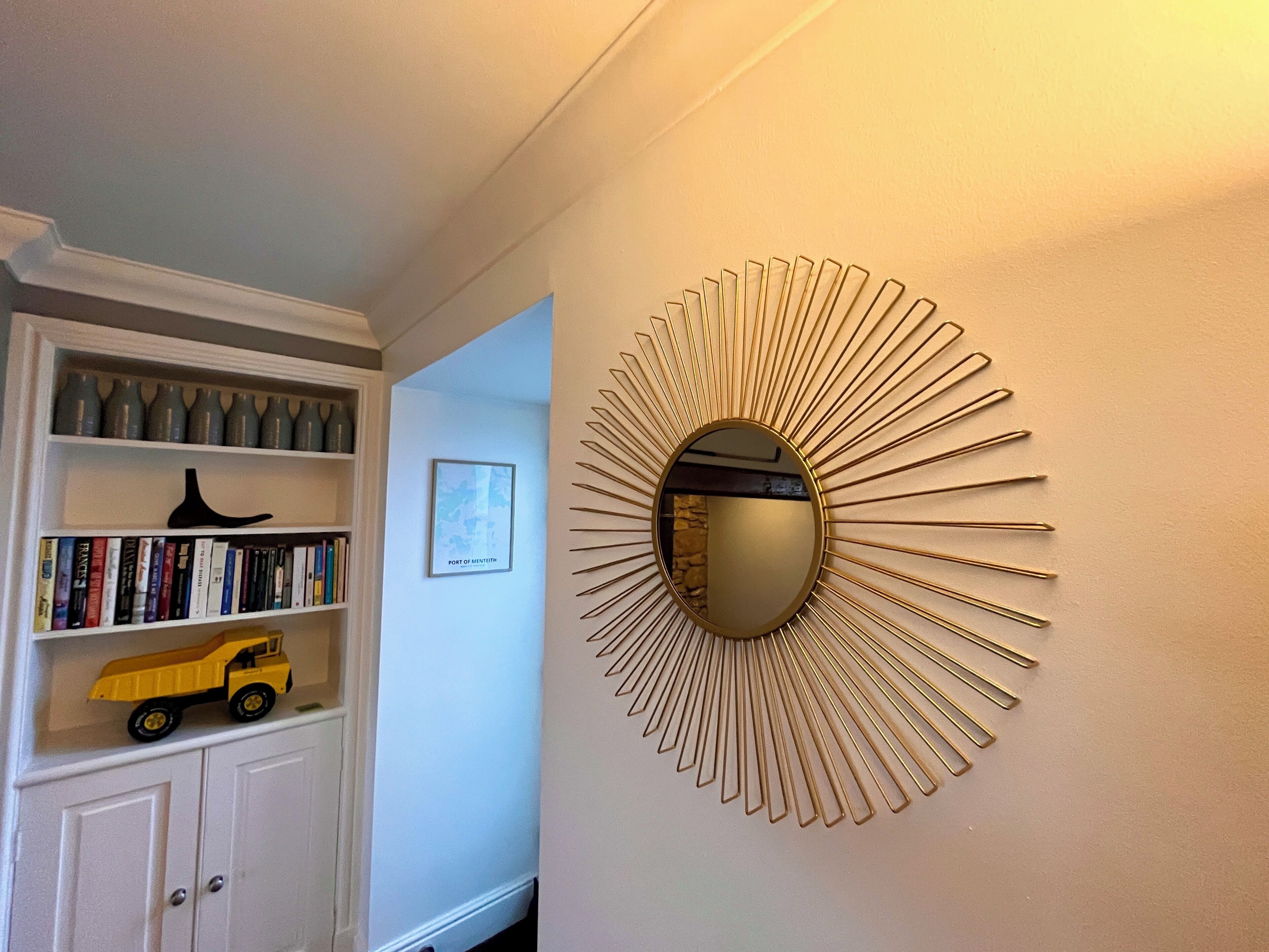 A hallway with a white cabinet containing books and a toy truck, a framed map on the wall, and a sunburst mirror.